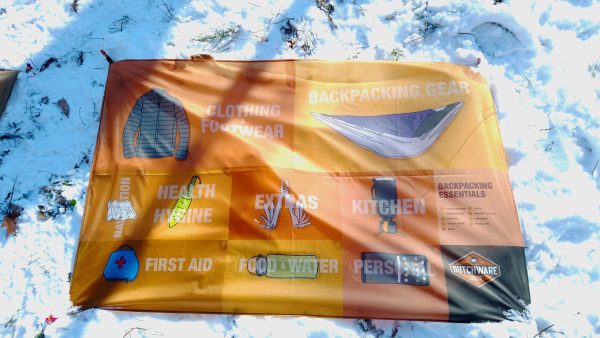 colorful poly fabric cloth with backpacking essential checklist prints on it laying flat on snowy grass